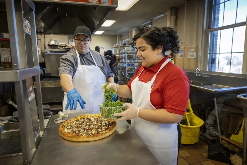 Tim Lopez, the culinary arts teacher, worked with Nataly Valentin, 17, right, as she spreads arugula onto a pizza during culinary arts class on Dec. 14, 2022.